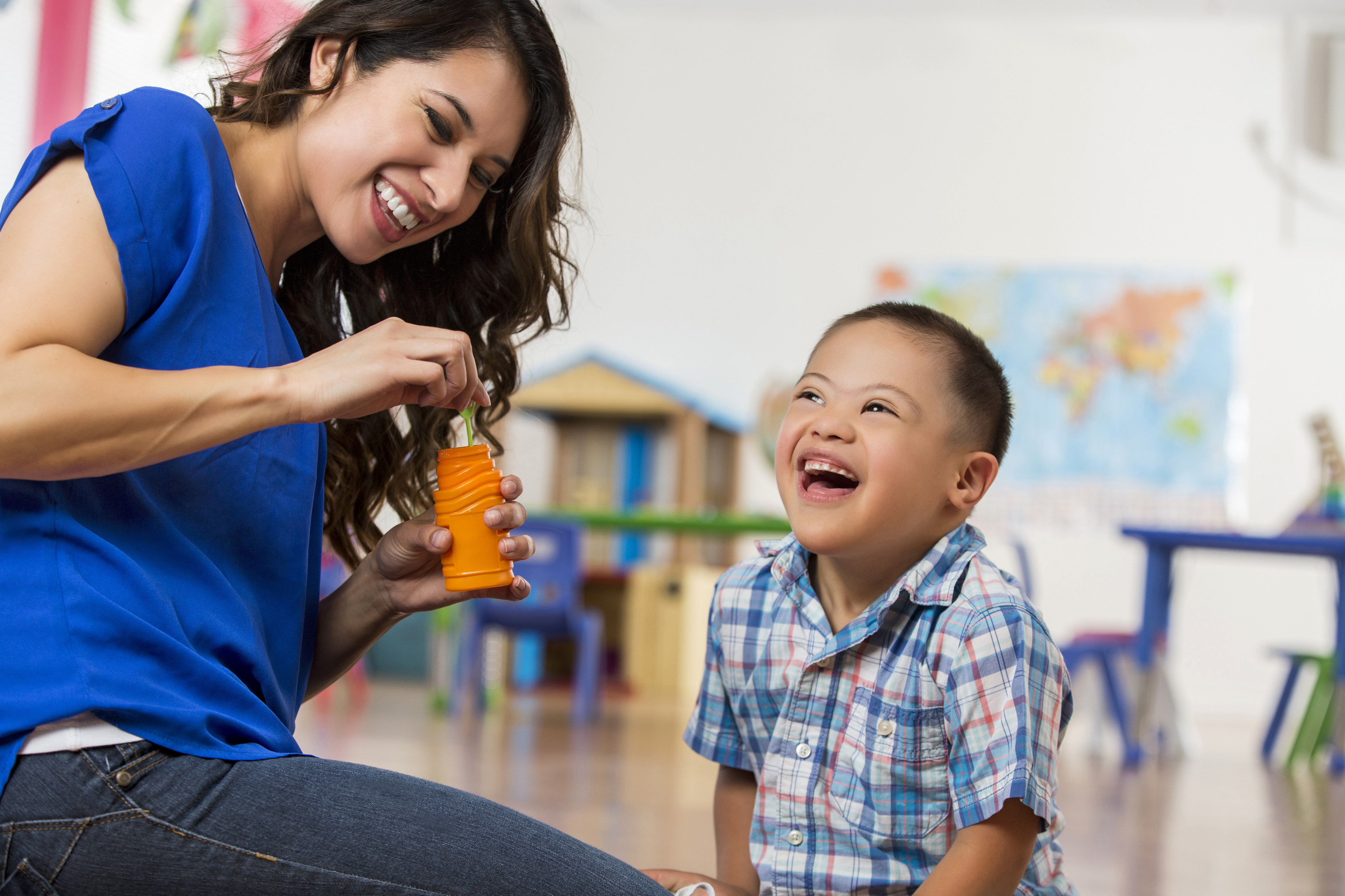 Picture of a woman holding a container of bubbles next to a boy (4-6 years old) in a classroom setting. Both people are smiling.